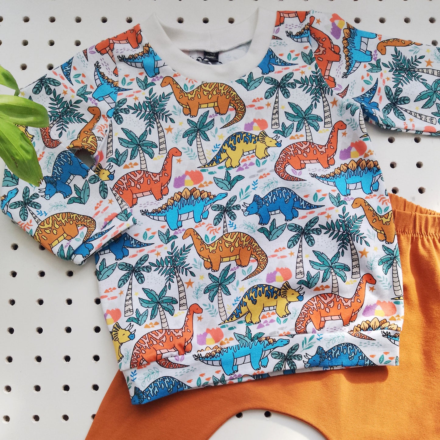 flatlay of a white long-sleeved childrens' t-shirt with colourful cartoon dinosaurs, and matching plain orange leggings