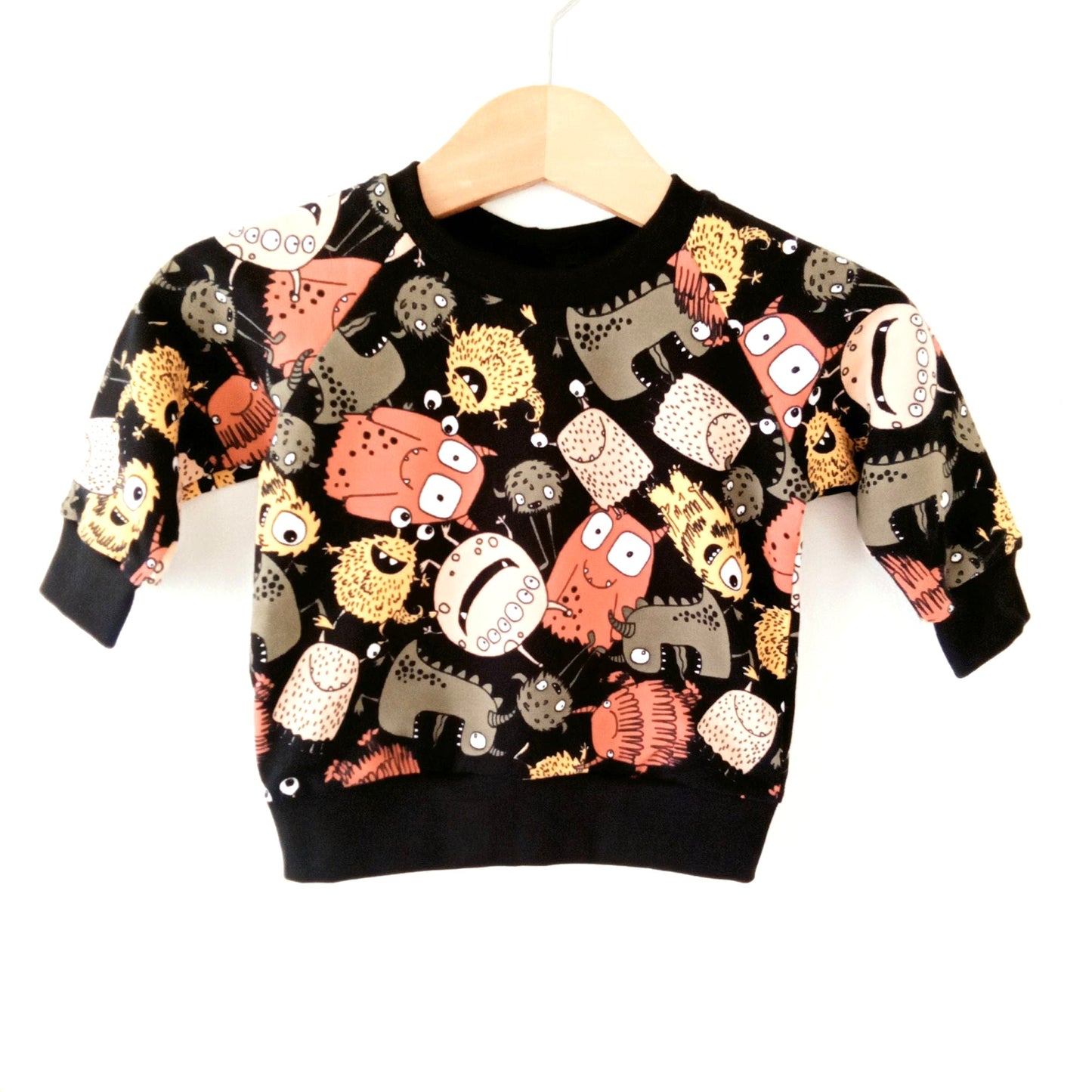 Still life of a black Khudu Kids baby sweatshirt, featuring colourful cartoon monsters and aliens.