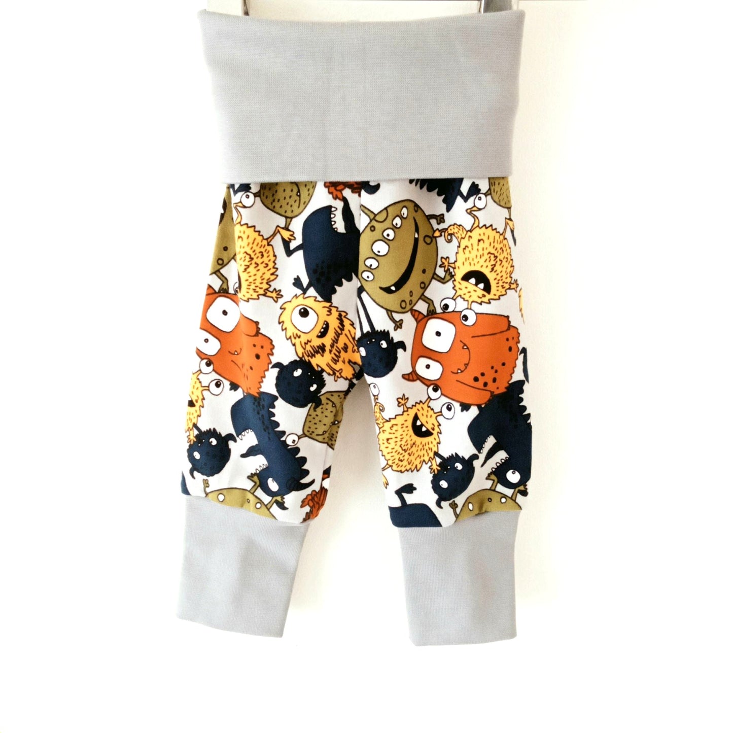 Still life of light grey Khudu Kids baby joggers with foldover waistband and ankle cuffs, featuring colourful cartoon monsters and aliens.